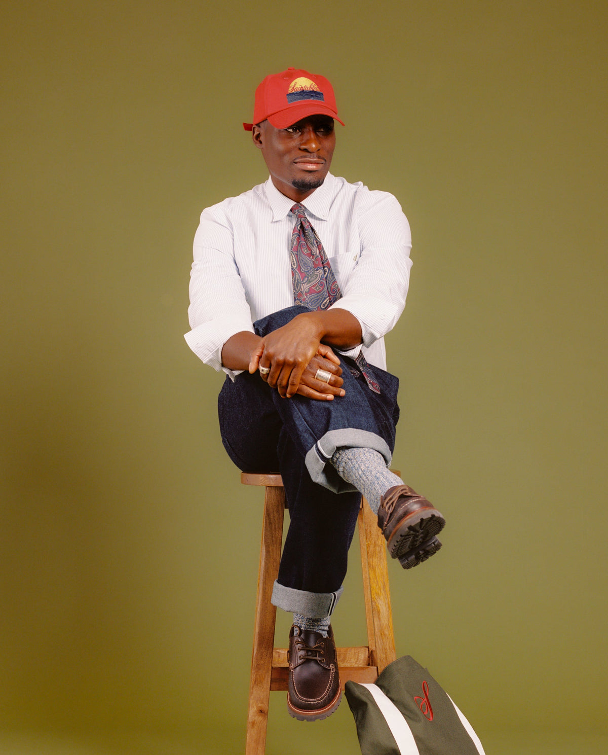A man wearing a red baseball cap, white shirt, patterned tie, dark trousers, and brown shoes sits on a wooden stool against an olive green studio background. A green and white bag rests on the floor beside the stool.